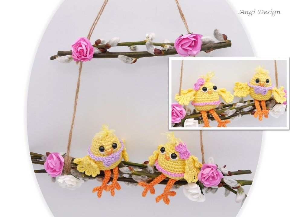 Handgehäkelte gelbe Küken mit lila Details auf einem Zweig, geschmückt mit Weidenkätzchen und rosa Stoffblüten vor hellem Hintergrund.