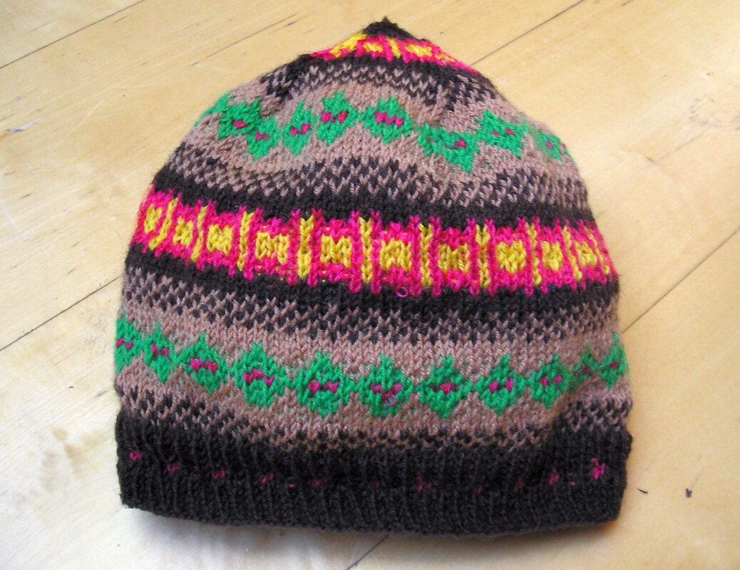 Gestrickte Beanie in Braun mit grünen, pinken und gelben Mustern auf hellem Holzuntergrund