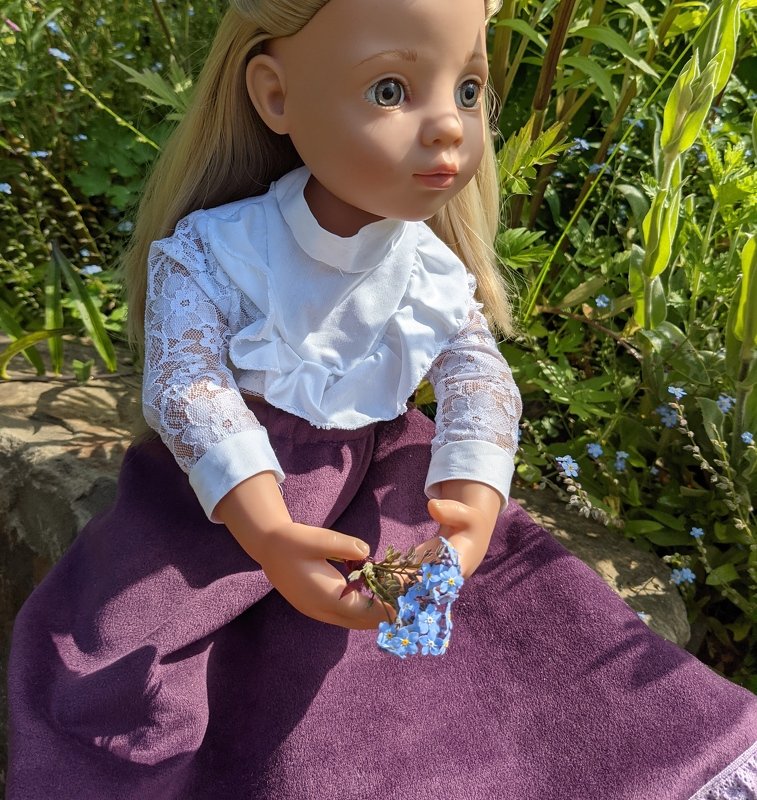 Nahaufnahme einer blonden Puppe mit weißer Bluse, transparenten Spitzenärmeln und lila Rock, sie hält kleine blaue Blumen