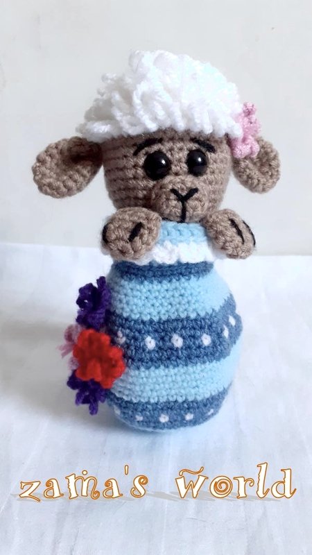 Gehäkeltes Lämmchen mit weißem Lockenkopf, braunem Gesicht, blau-weiß gestreiftem Kleid und bunten Häkelblumen