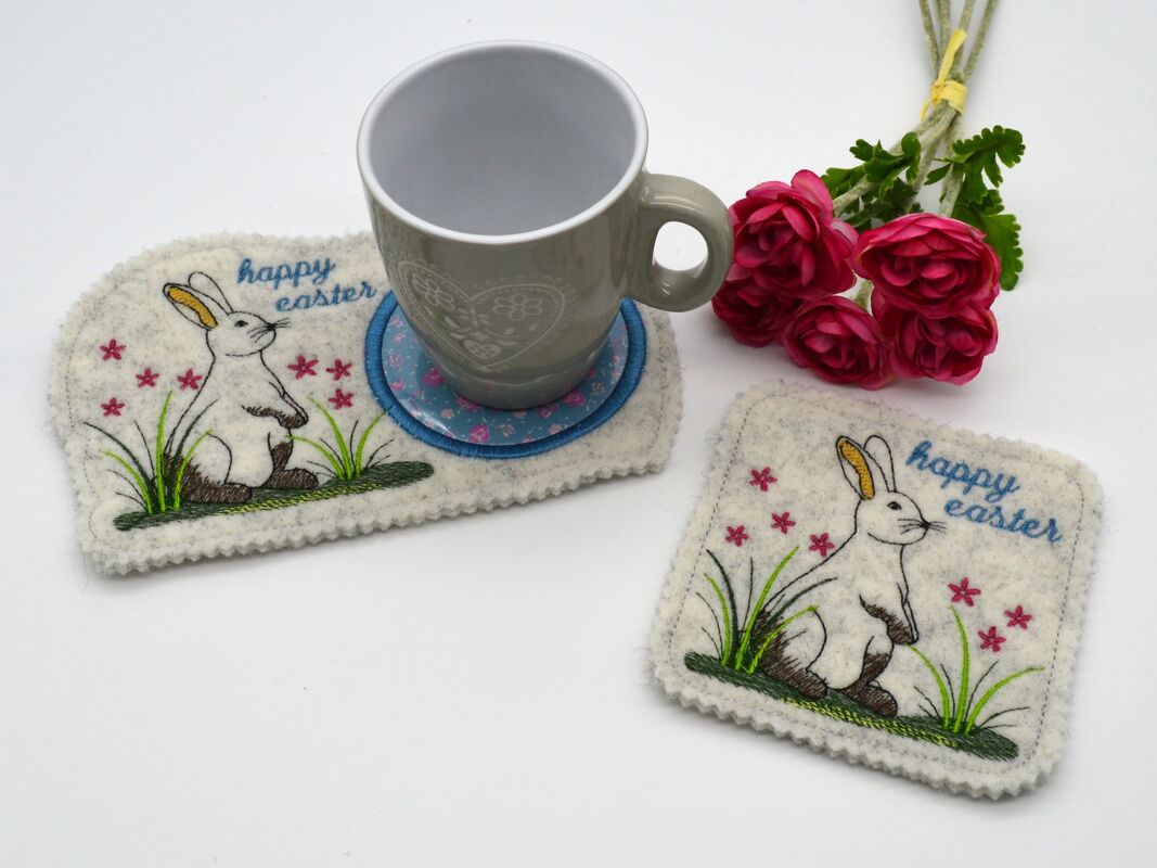 Filzuntersetzer mit gesticktem Hasen, Gras und kleinen pinken Blüten; eine graue Tasse steht auf einem Untersetzer, daneben ein kleiner Blumenstrauß.