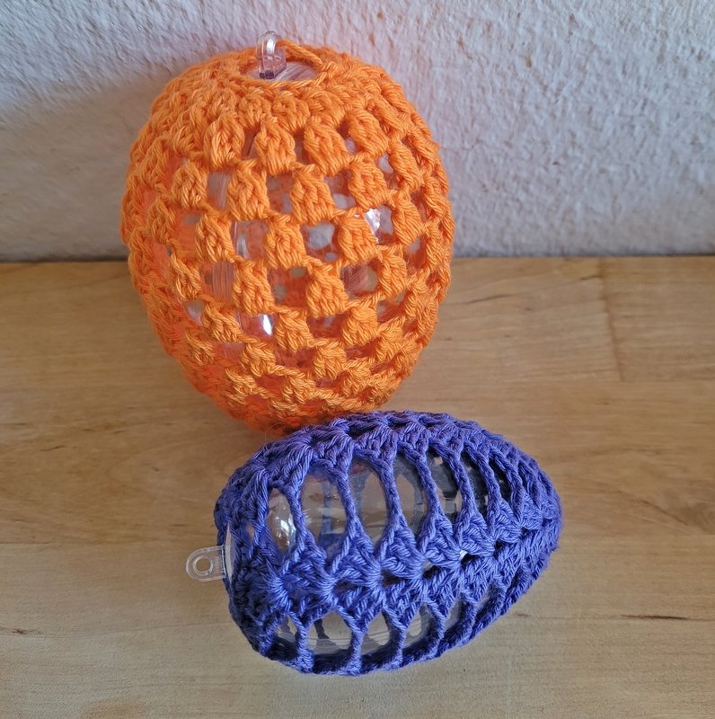 Zwei gehäkelte Ostereier, eins orange, eins lila, mit transparenten Einsätzen und Aufhängern auf Holzuntergrund vor heller Wand.