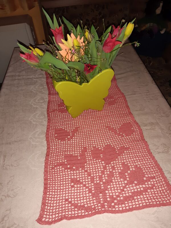Frühlingshaft gedeckter Tisch mit lachsfarbenem gehäkeltem Läufer und buntem Tulpenstrauß in gelber Schmetterlingsvase.