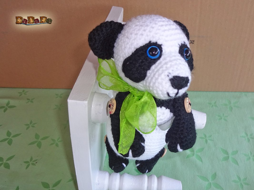 Gehäkelter Panda (schwarz-weiß) mit blauen Augen und grünem Band auf weißer Halterung