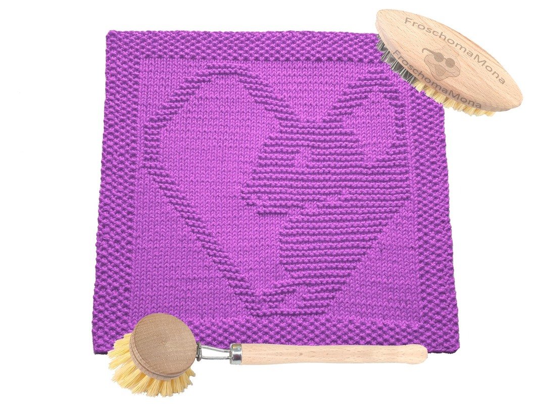 Lila gestricktes quadratisches Tuch mit eingearbeitetem Herzmotiv und zwei hölzernen Bürsten auf weißem Hintergrund.