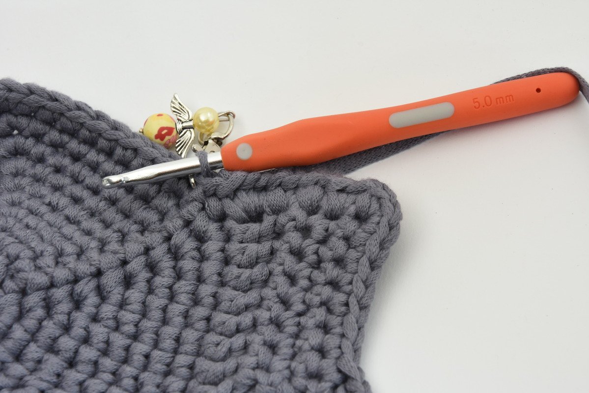 Gehäkelte graue Fläche mit orangefarbener Häkelnadel 5,0 mm und Perlen-Maschenmarkierer