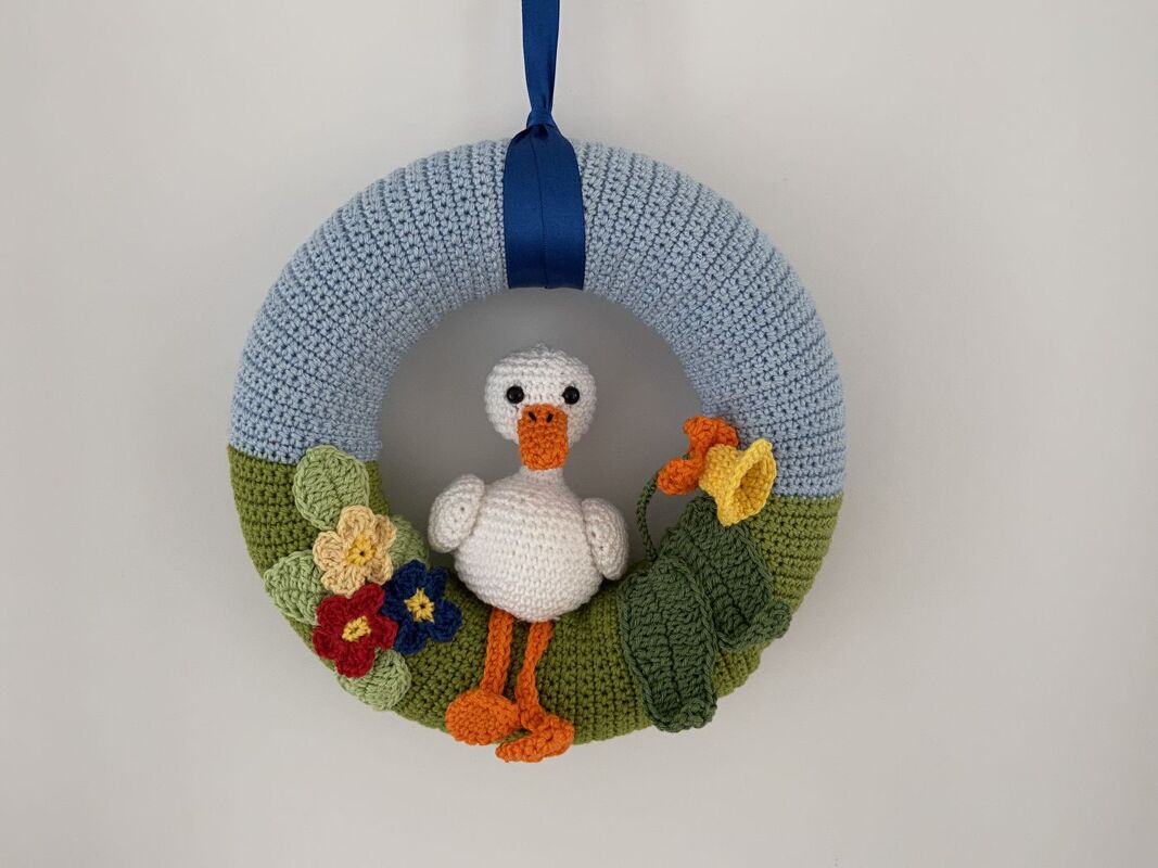 Gehäkelter Türkranz in Hellblau und Grün mit kleiner weißer Ente, bunten Häkelblüten, grünen Blättern und blauem Aufhängeband.