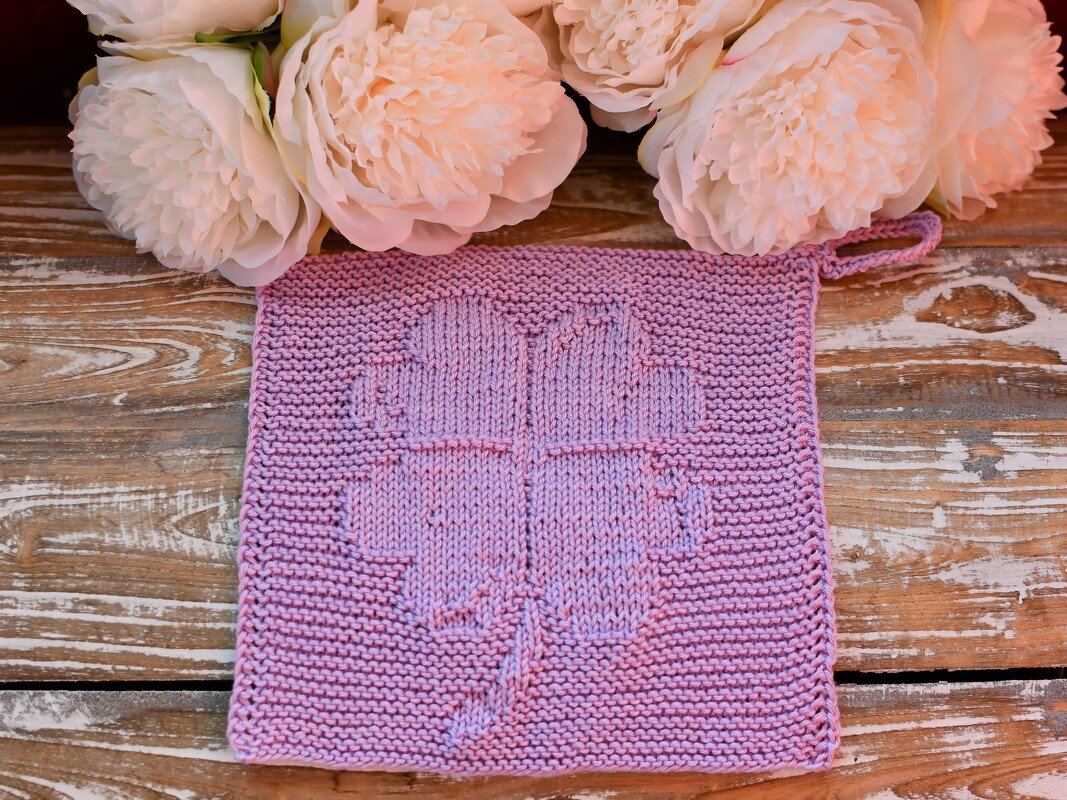 Lilac hand-knitted square with a raised four-leaf clover and hanging loop on a wooden surface with pale flowers.