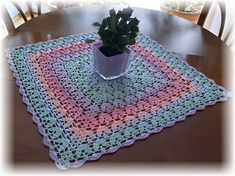 Quadratisches gehäkeltes Deckchen in Pastellfarben mit Muschelmuster und Spitzenrand auf einem Holztisch, mit einer Topfpflanze in der Mitte.