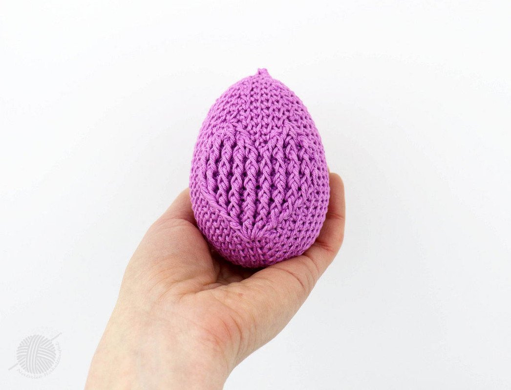 Hand hält ein lila gehäkeltes Ei mit Zopfmuster vor weißem Hintergrund.