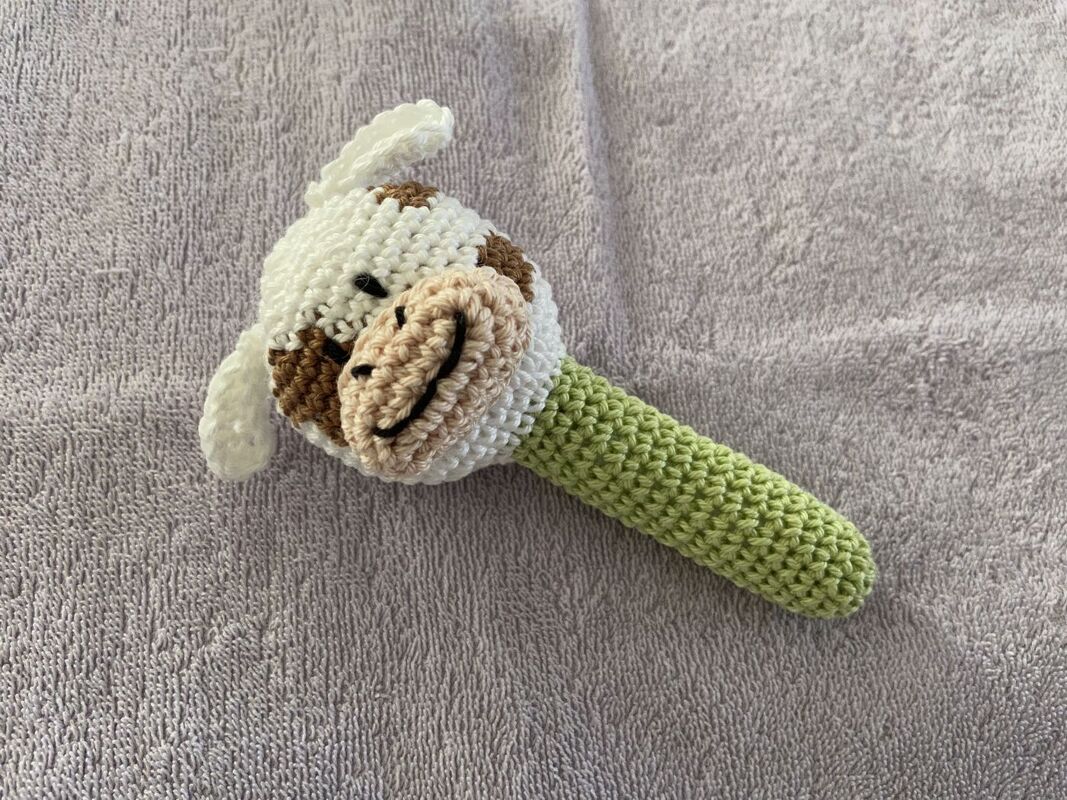 Gehäkelte Babyrassel in Kuhform mit grünem Griff auf grauem Handtuch.