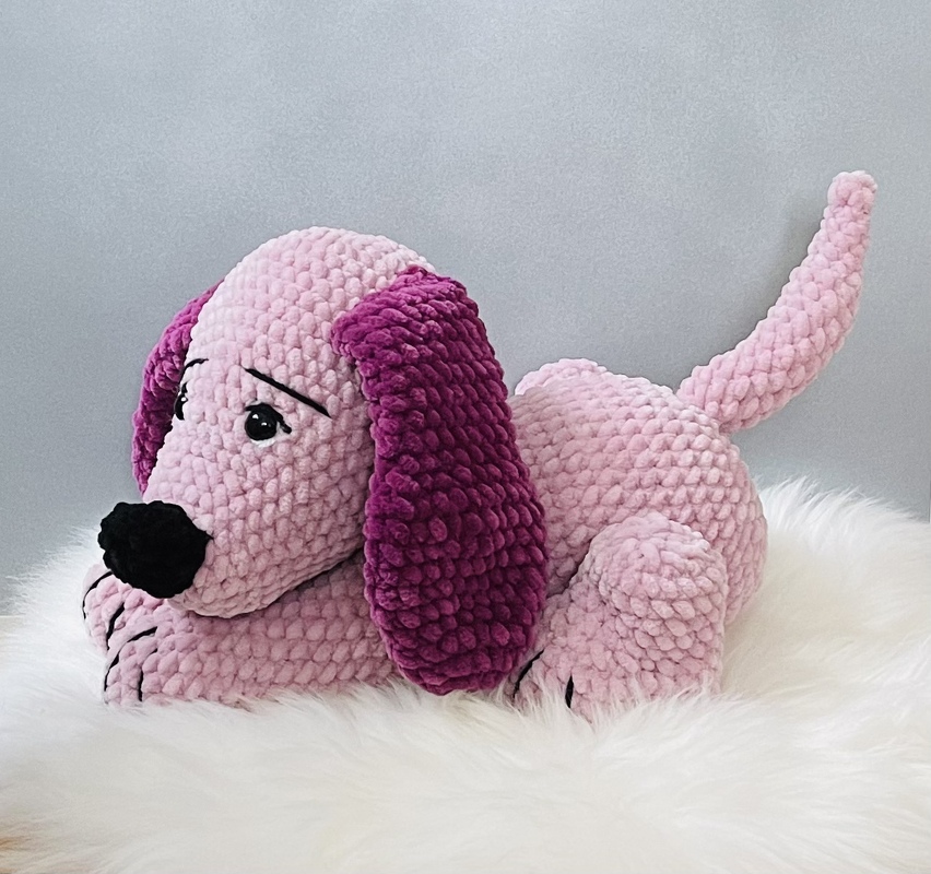 Rosa gehäkelter Amigurumi-Hund mit dunkelpinken Ohren liegend auf weißem Fell