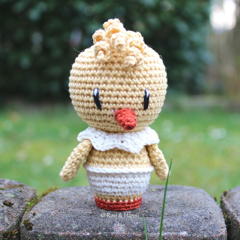 Gehäkeltes Amigurumi-Küken in Hellgelb mit weißem Kragen und orangefarbenem Schnabel, stehend auf einem Stein.