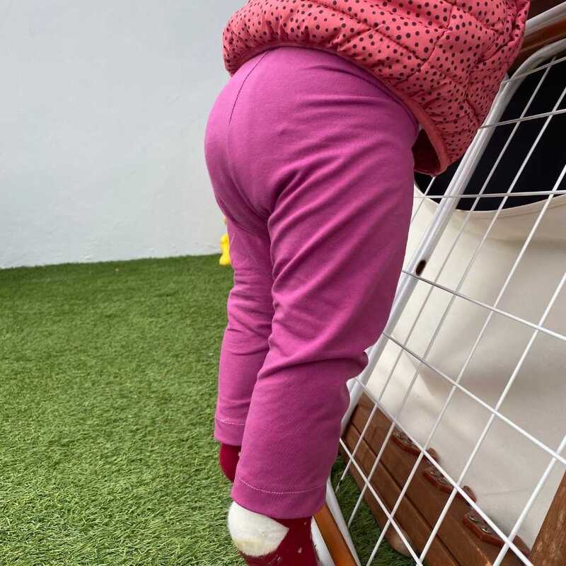 Kleinkind von hinten in pinken Leggings und gepunkteter Jacke, klettert an einem weißen Gitter auf grünem Kunstrasen.