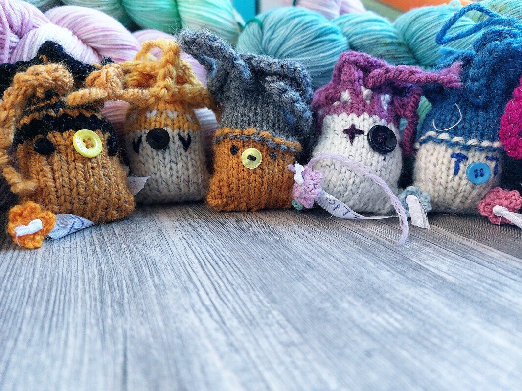 Sechs handgestrickte Amigurumi-Säckchen in verschiedenen Farben auf einer Holzfläche, mit Garnknäueln im Hintergrund.