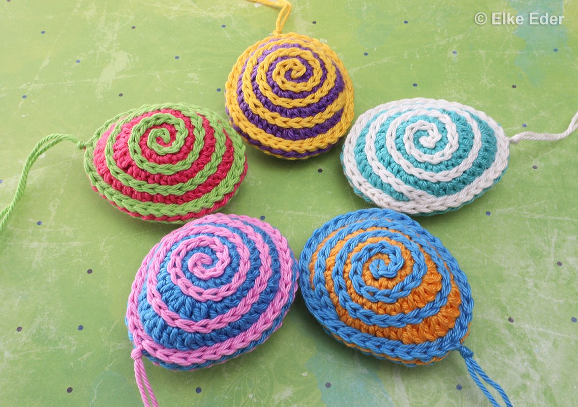 Five colorful crocheted spiral eggs with hanging loops on a green background