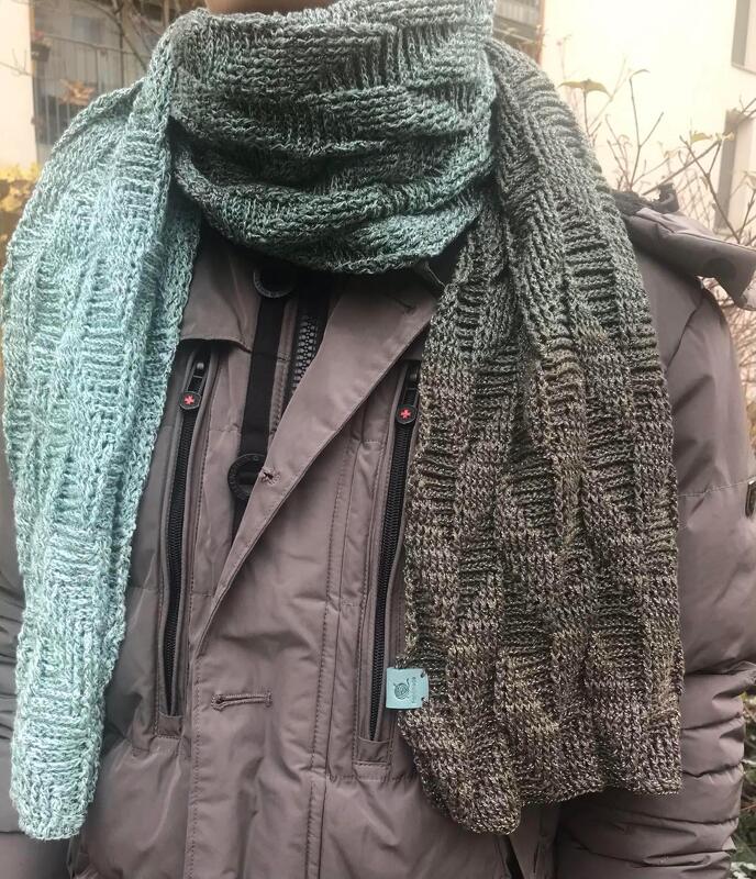 Person trägt einen gehäkelten Schal mit Farbverlauf in Grün- und dunklen Grüntönen und Reliefstruktur über einer grauen Jacke.