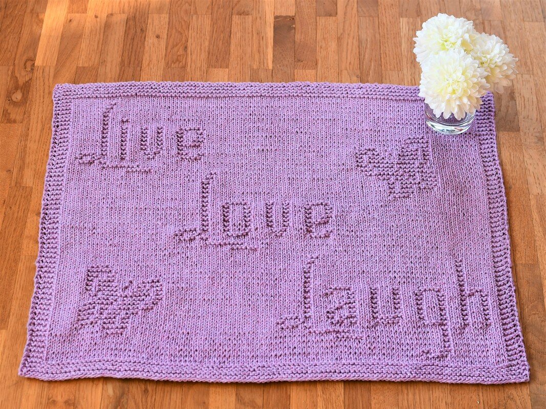 Rectangular purple knitted mat with 'Live Love Laugh' text and heart motifs on a wooden floor beside a small glass of white flowers.