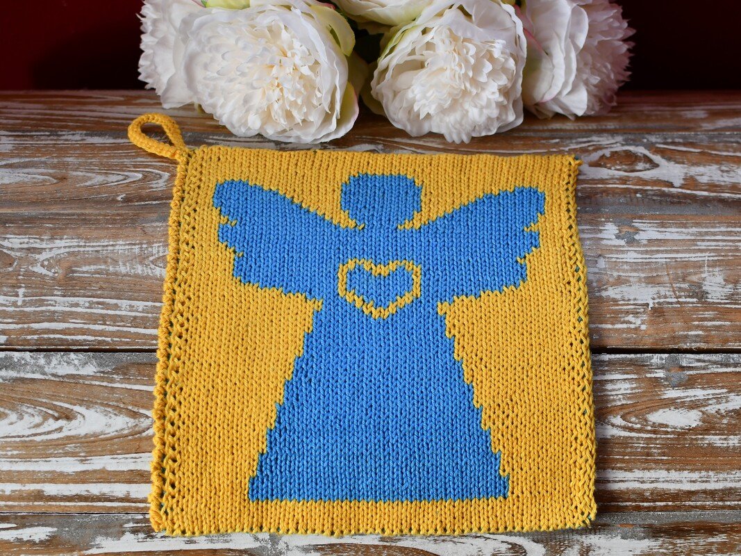 Quadratischer gelber Topflappen mit blauem Engelmotiv und Herz auf rustikalem Holztisch, oben wei Blumen.