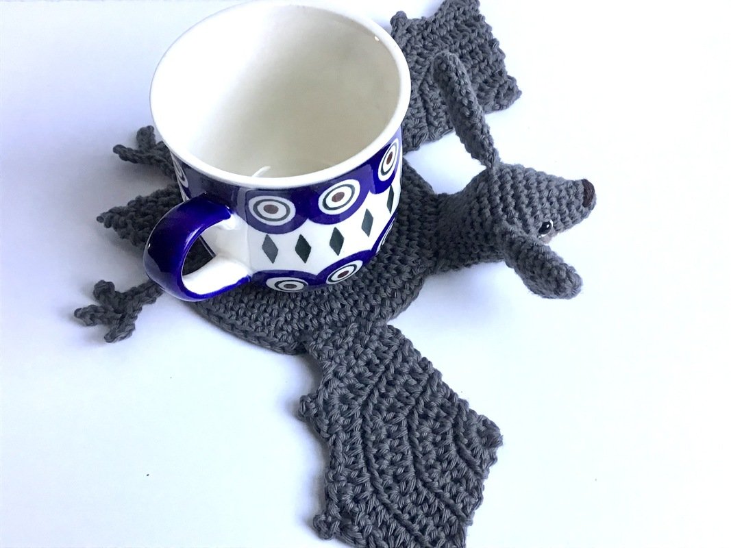 Dunkelgrauer gehäkelter Fledermaus-Untersetzer unter einer weißen Tasse mit blauem Muster.