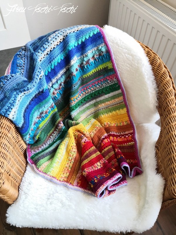 Bunte gehäkelte Decke mit Regenbogenverlauf auf einem Korbstuhl mit weißem Fellsitz