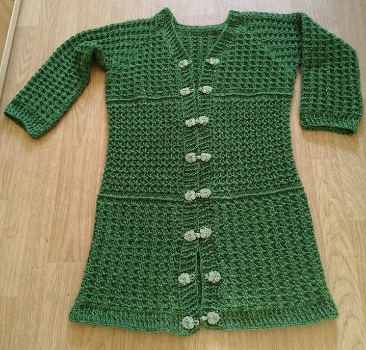 Langärmelige dunkelgrüne Häkeljacke mit V‑Ausschnitt und hellen Blütenverschlüssen auf Holzfußboden.