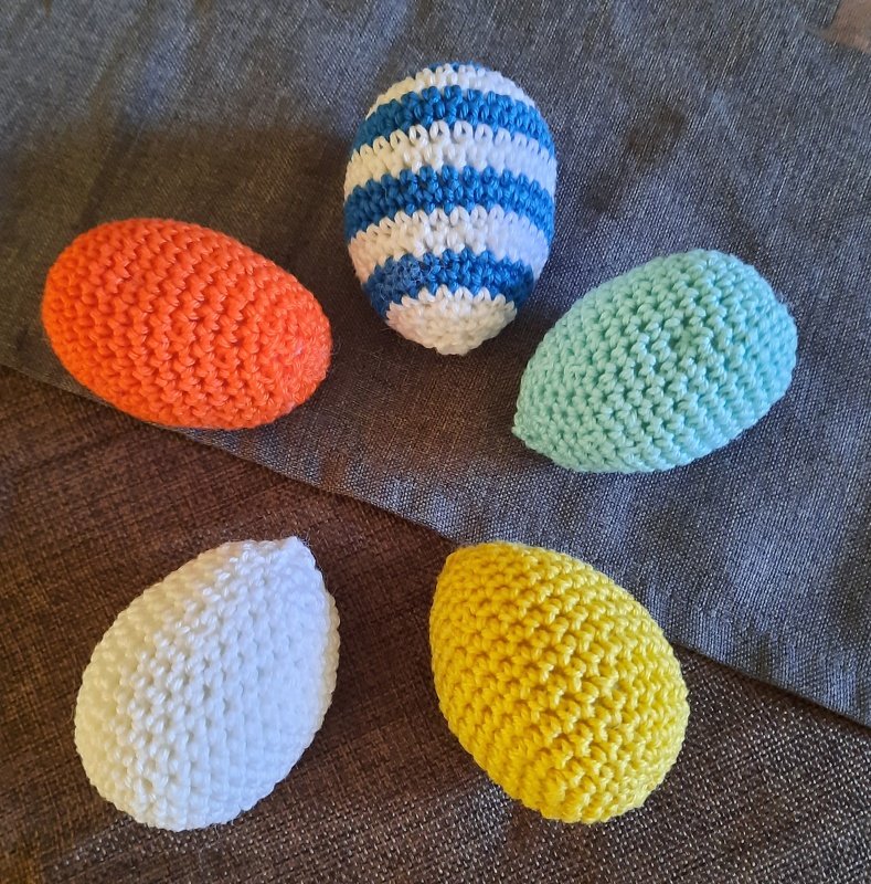 Fünf handgehäkelte Ostereier in Orange, Blau-Weiß gestreift, Hellblau, Weiß und Gelb auf dunklem Stoff.