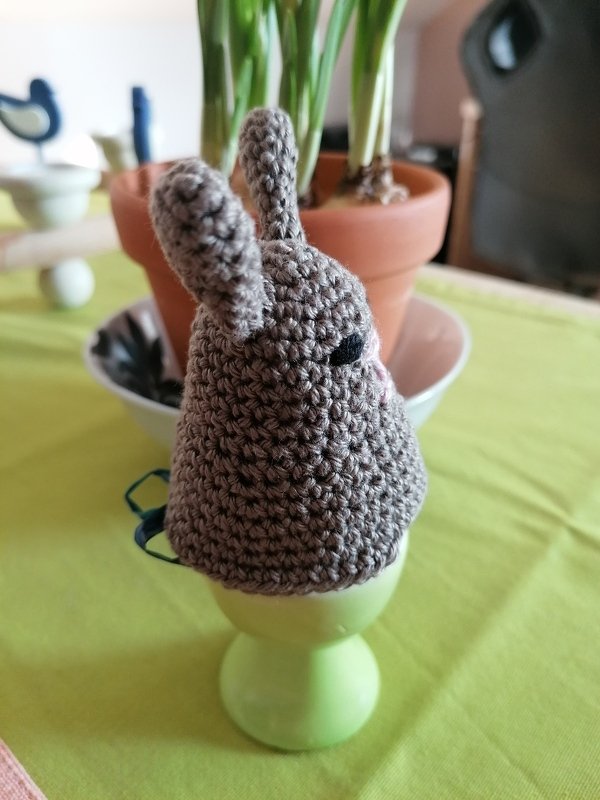 Gehäkelter Eierwärmer in Hasenform (grau-braun) auf hellgrünem Eierbecher, Topfpflanze im Hintergrund