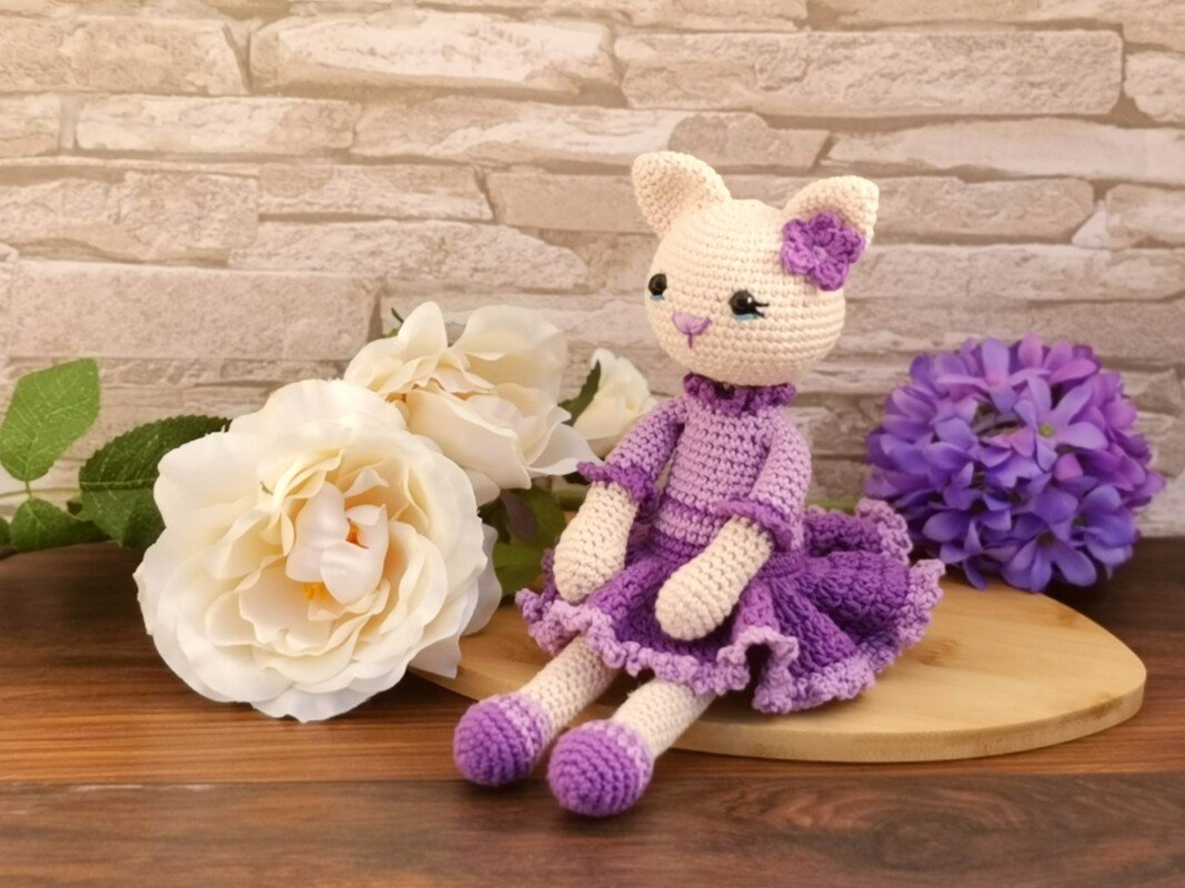 Cream and purple crocheted amigurumi cat doll seated beside white roses and a purple hydrangea