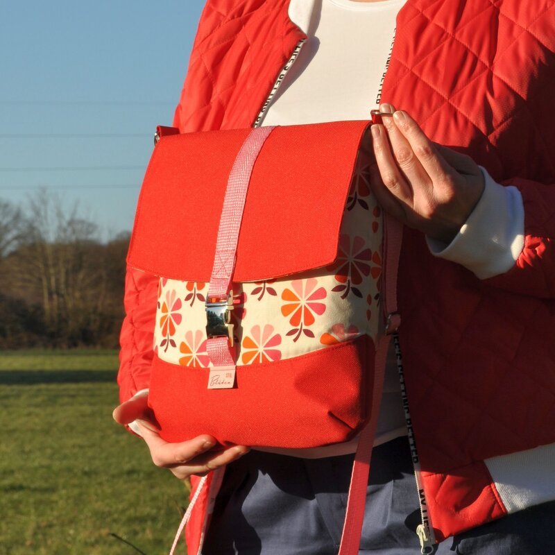 Person in roter Steppjacke hält eine rote Stofftasche mit floralem Muster und Schnallenverschluss im Freien.
