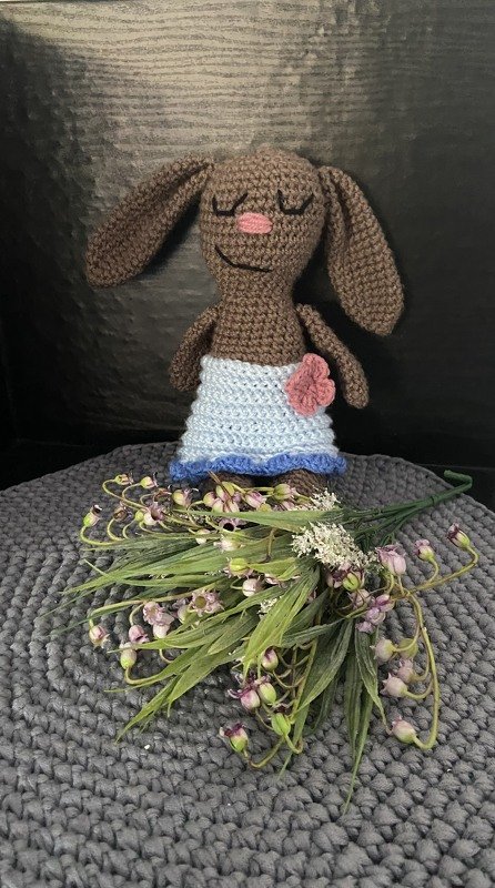 Brauner gehäkelter Hase mit geschlossenen Augen und rosa Nase, trägt hellblaues Kleid mit rosafarbener Blüte, sitzt auf grauem gehäkeltem Untergrund vor Kunstblumen.