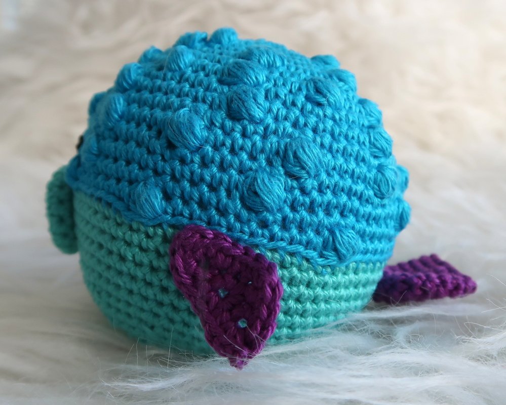 Kugelförmiges blau-türkis gehäkeltes Amigurumi mit Noppenmaschen und lila Flossen