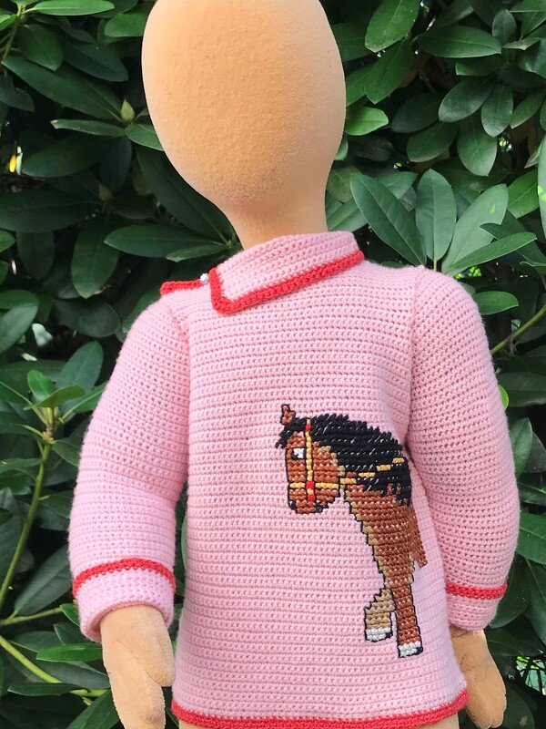 Rosa gehäkelter Kinderpullover mit rotem Rand und Pferde-Applikation auf Schaufensterpuppe