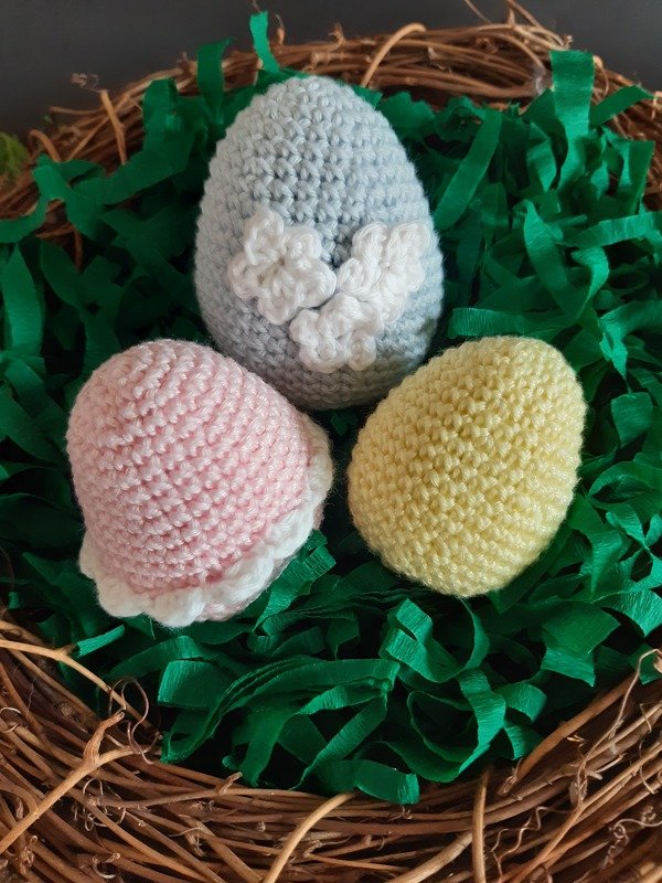 Drei gehäkelte Eier: hellblau mit kleinen Blüten, rosa mit Rüsche, gelb glatt, platziert in Weidenkranz mit grünem Papiergras