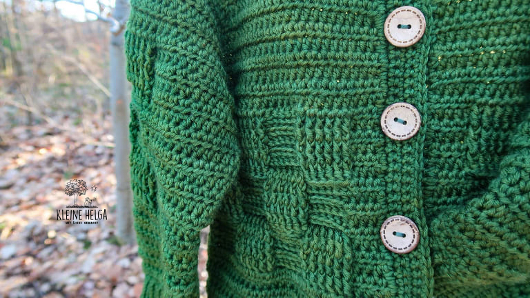 Nahaufnahme einer grünen gehäkelten Kinderjacke mit drei hellen Holzknöpfen vor unscharfem Waldhintergrund.