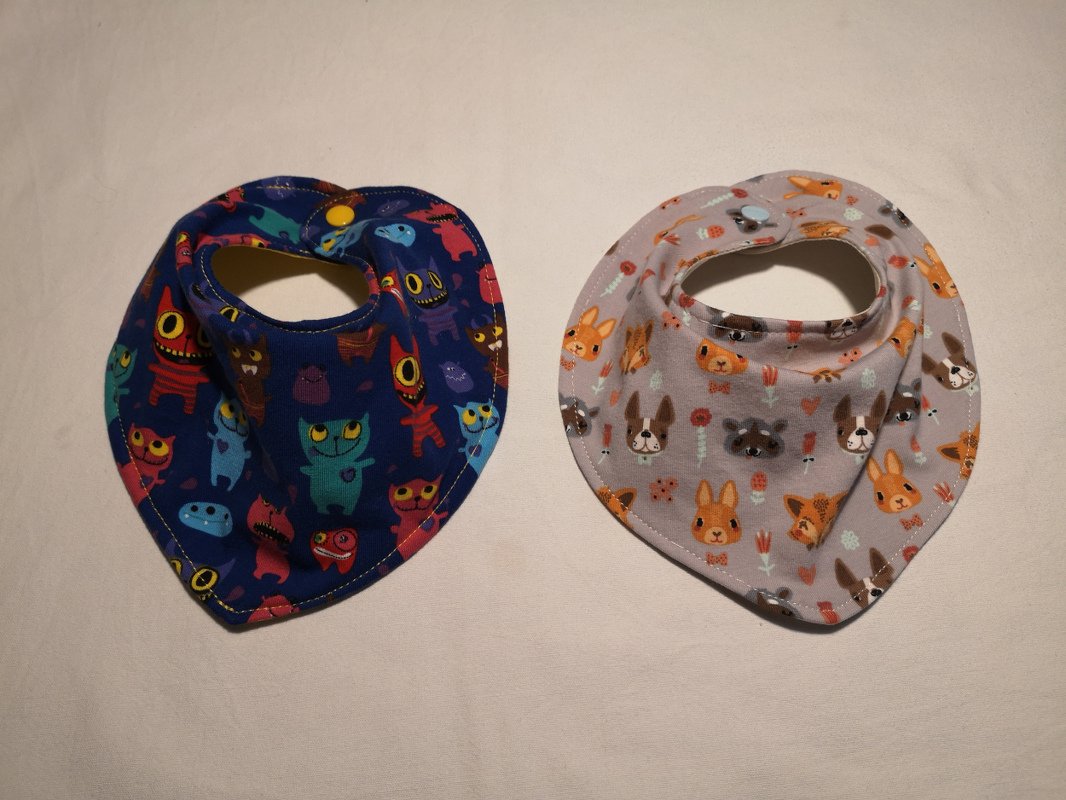 Zwei Bandana-Halstücher nebeneinander: links blaues mit Monster-Motiv, rechts hellgraues mit Tiermotiven, beide mit Druckknopf