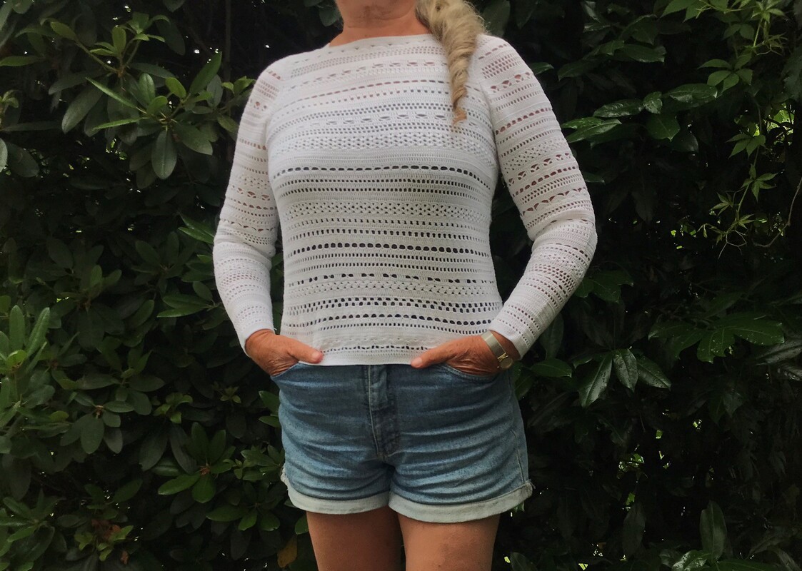 Frau trägt weißes Langarm-Häkelshirt mit durchbrochenen Mustern und Jeansshorts vor grünem Blattwerk.