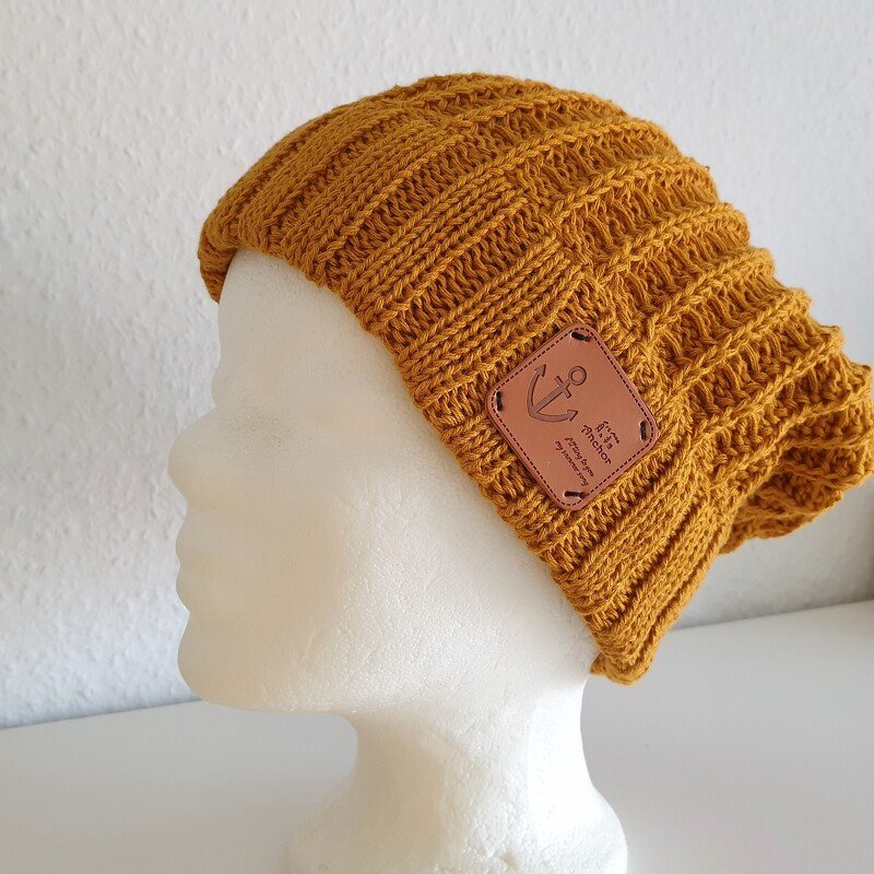 Senfgelbe gestrickte Mütze mit geripptem Muster und braunem Anker-Patch auf weißem Puppenkopf