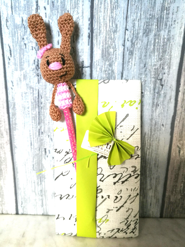 Brauner gehäkelter Amigurumi-Hase mit rosa Schleife als Stift-Topper vor verpacktem Geschenk mit grünem Band