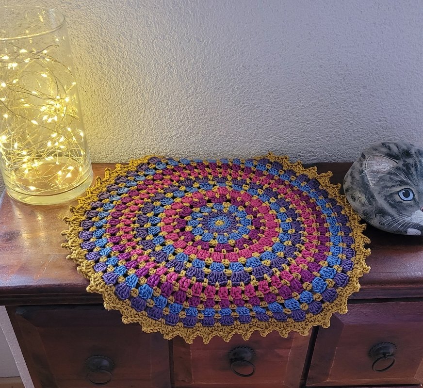 Rundes gehäkeltes Deckchen in mehreren Farben auf dunklem Holz, links Glas mit Lichterkette, rechts Katzendeko