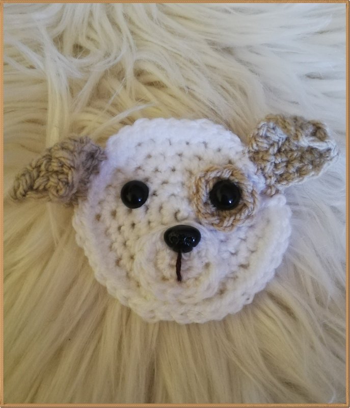 Nahaufnahme einer gehäkelten Hundeapplikation in Weiß und Beige mit schwarzen Augen und Nase auf flauschigem Hintergrund