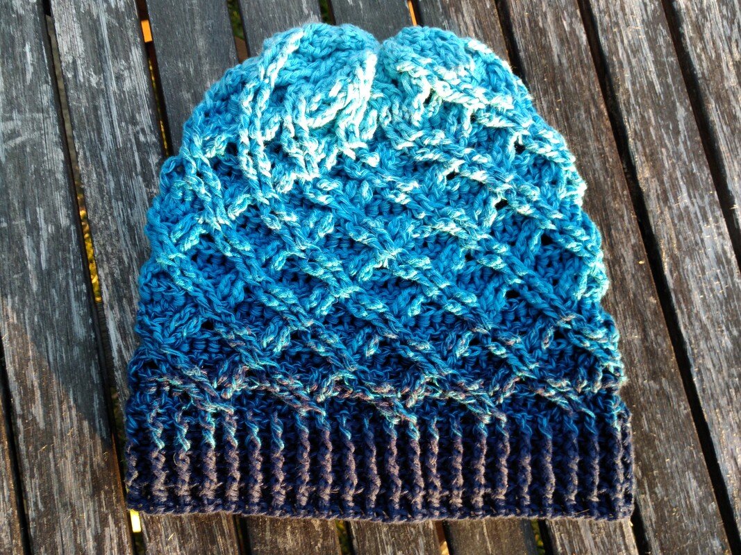 Gehäkelte Damenmütze in Blau mit Farbverlauf und Rippbündchen, liegend auf Holzdielen