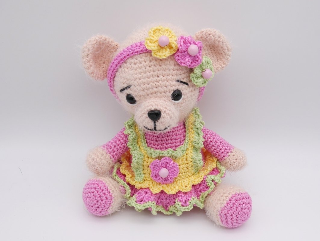 Gehäkelter Amigurumi-Teddybär in Rosa mit gelb-rosa Rüschenkleid und Blumenstirnband, sitzend.