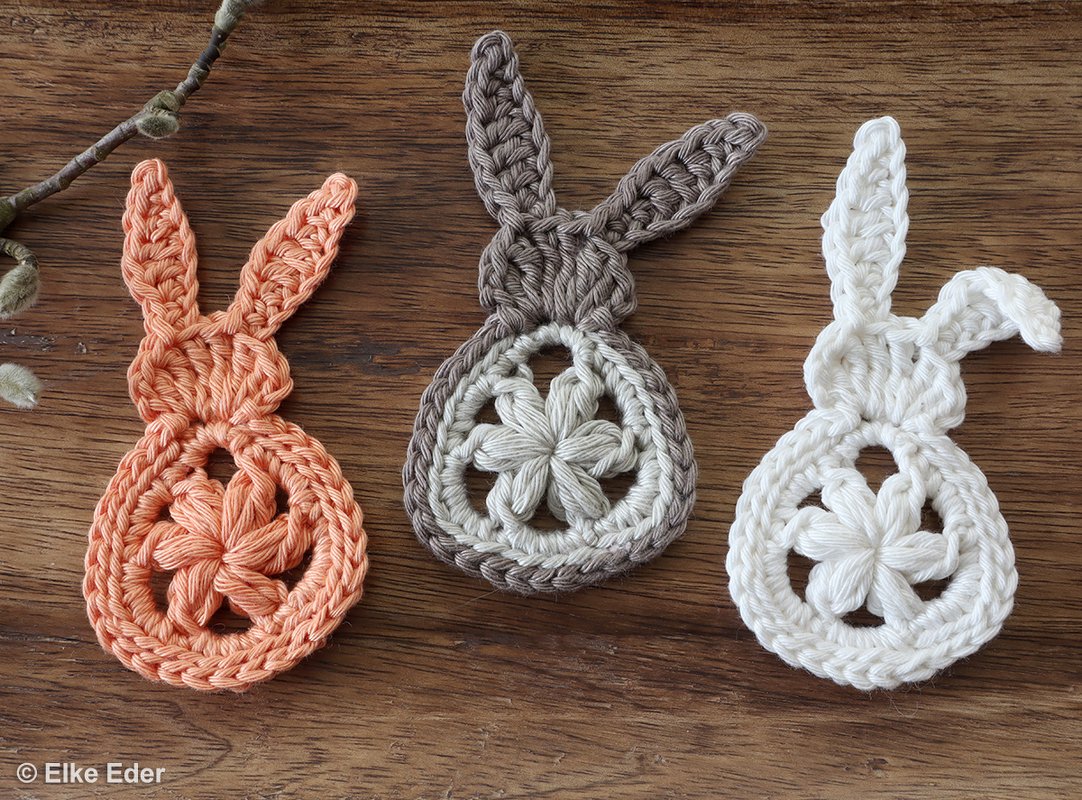 Three crocheted bunny motifs in orange, gray, and white with flower centers on a wooden surface.