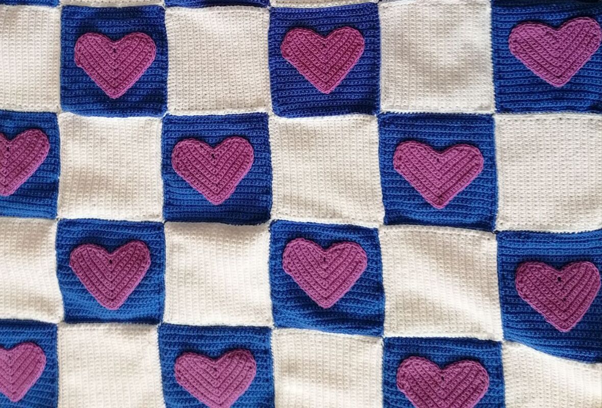 Gehäkelte Babydecke mit blau-weißem Schachbrettmuster und lila Herzapplikationen auf den blauen Feldern