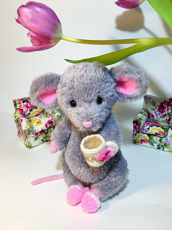Grey amigurumi mouse with pink ears and feet holding a small crochet cup, floral boxes and pink tulip in background