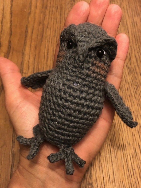 Nahaufnahme einer handgroßen, grau gehäkelten Amigurumi-Eule mit sichtbaren Maschen und Flügeln