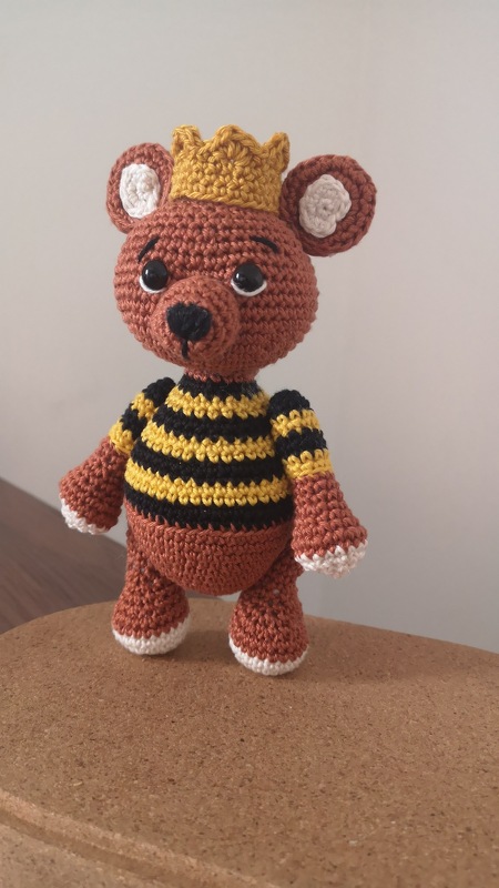 Gehäkelter Teddybär mit gelber Krone und schwarz-gelbem Ringelshirt auf Korkuntergrund