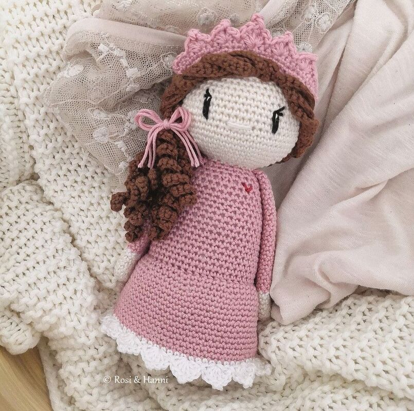 Gehäkelte Amigurumi-Puppe mit rosa Kleid, weißem Spitzenrand, braunen lockigen Haaren und rosa Krone auf einer gestrickten Decke