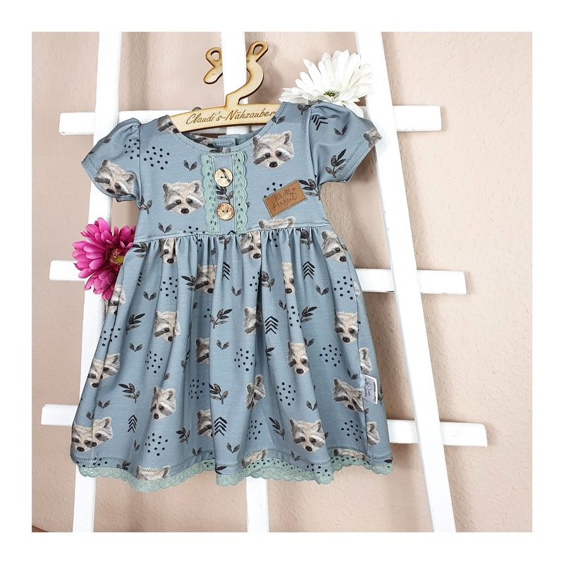 Hellblaues Kinderkleid mit Waschbär-Print, Spitzenbesatz und Knopfleiste, an einem Bügel vor einer weißen Leiter hängend.